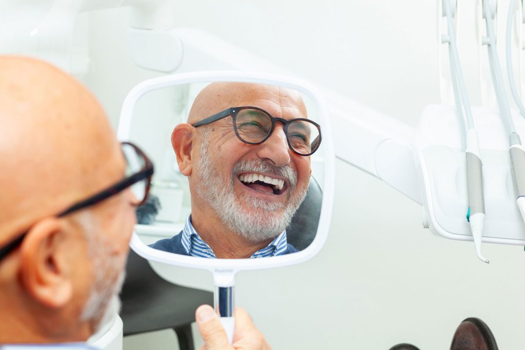 man smiling while holding a handheld mirror in a dental chair